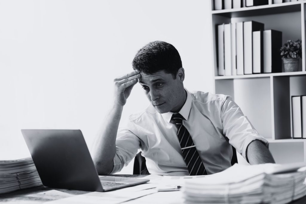 Frustrated professional man in office looking at laptop, showing signs of IT support issues.