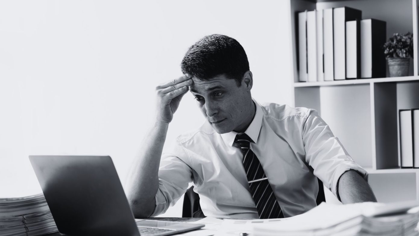 Frustrated professional man in office looking at laptop, showing signs of IT support issues.